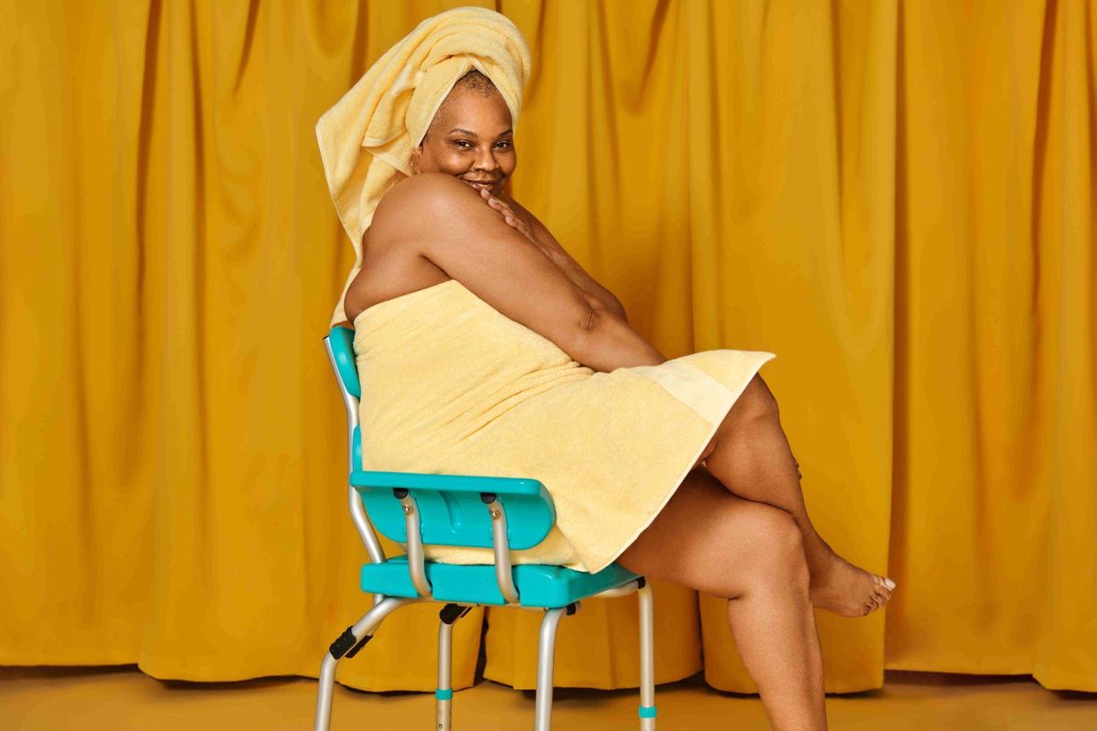 A smiling woman wrapped in a yellow towel on the teal Bue Shower Chair